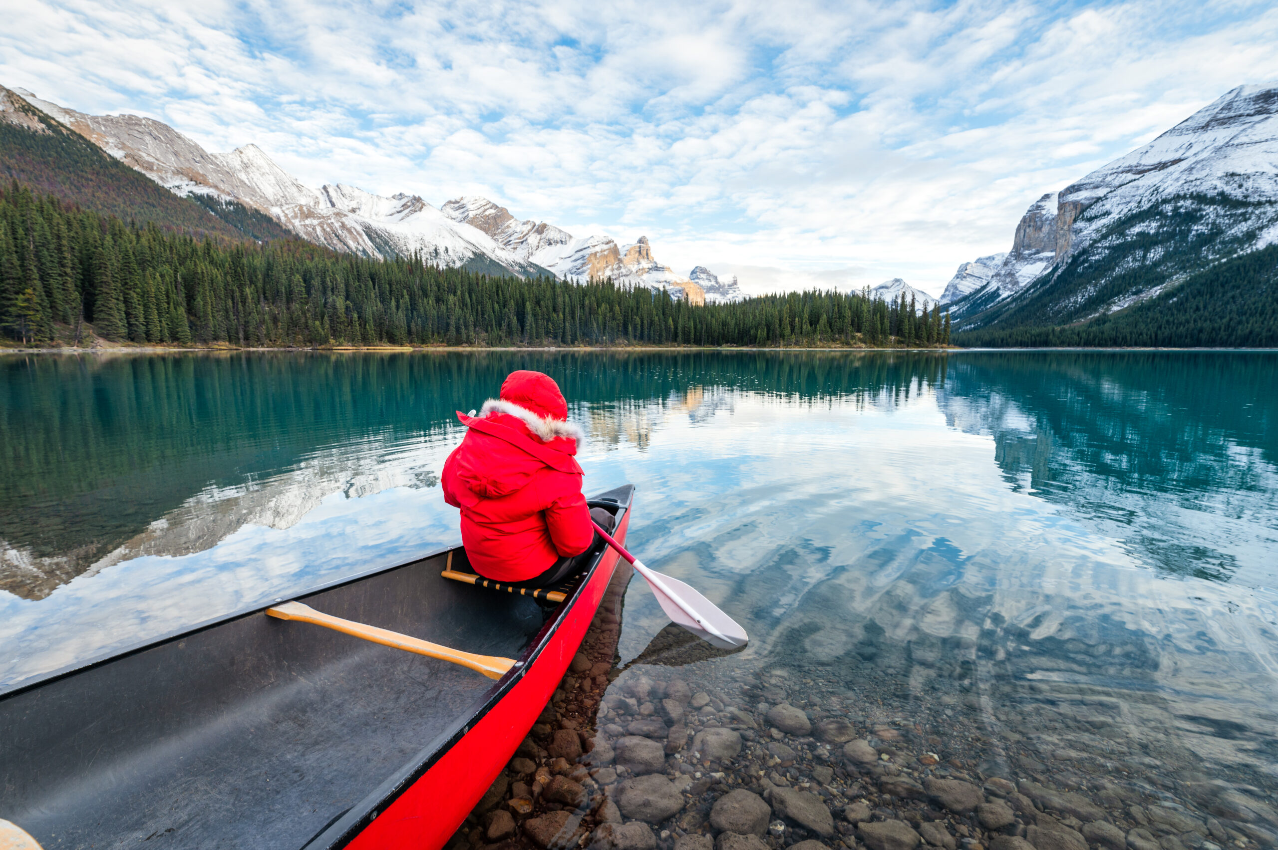 Child in red coat paddling canoe on Vancouver lake with snowy mountains in background.